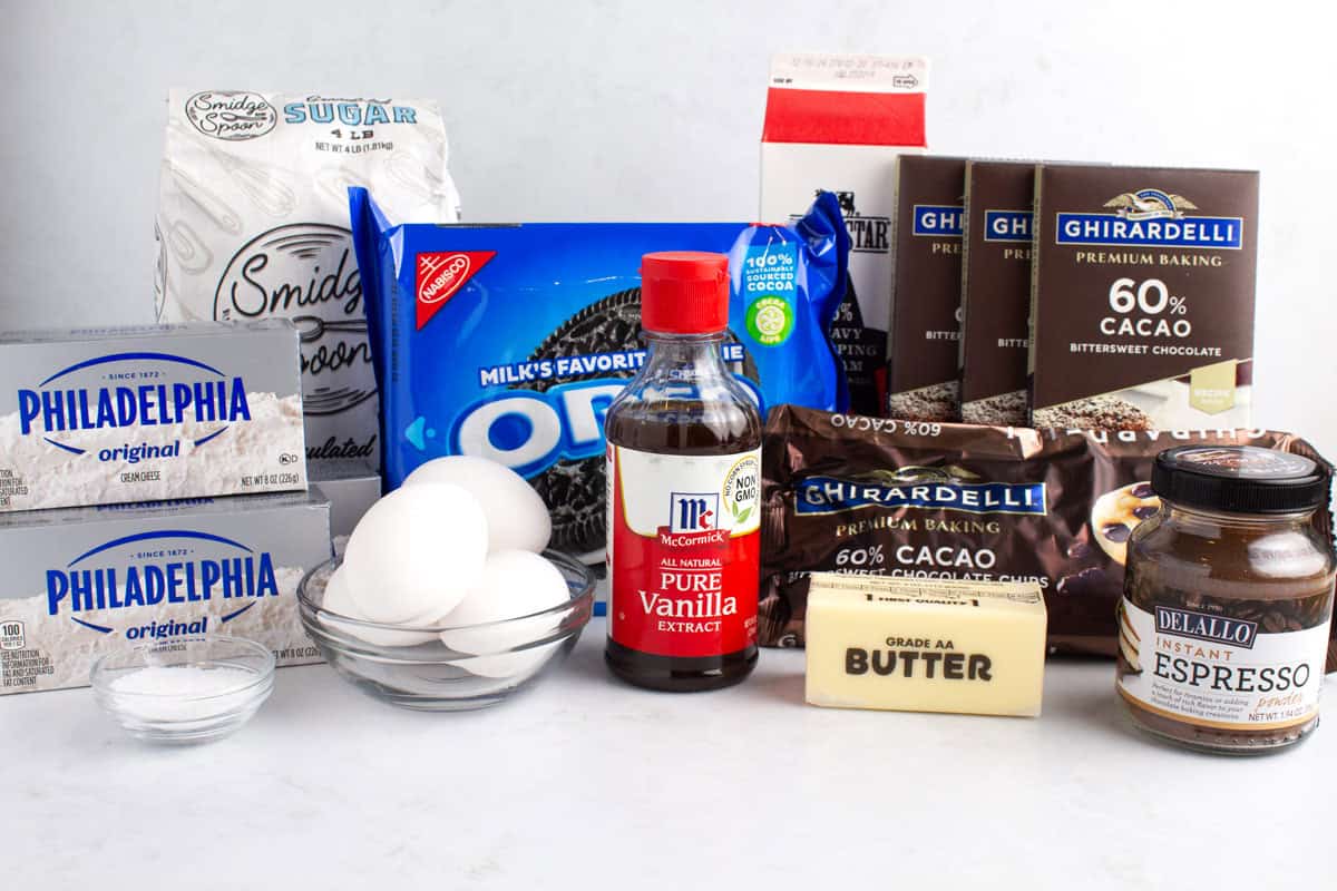 Two boxes of cream cheese, sugar, eggs, salt, Oreos, vanilla extract, a stick of butter, espresso, chocolate chips, milk, and bittersweet chocolate sitting on a white countertop.