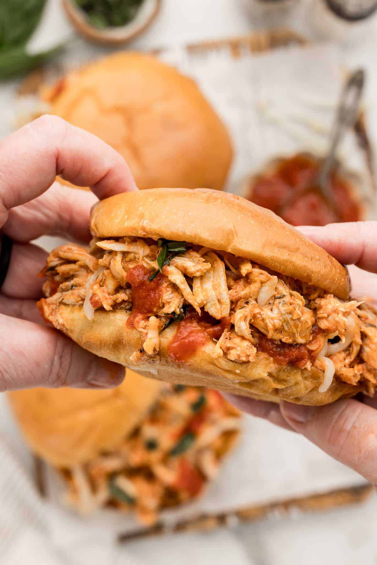 A pair of hands holding a shredded chicken parmesan sandwich over a table with two more sandwiches and two bowls of sauce and herbs on it.