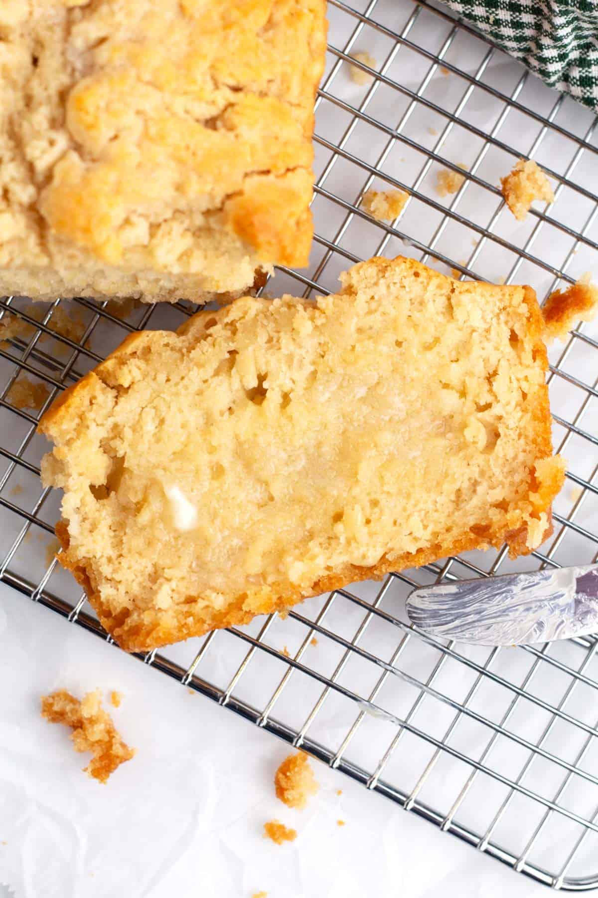 A slice of buttered bread on a wire rack with a butter knife on the side and a full loaf of the bread in the left corner.