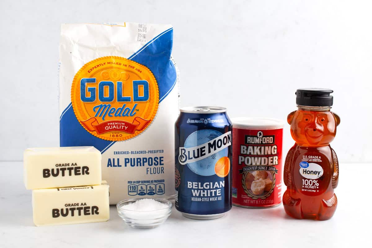 Flour, butter, salt, baking powder, a can of beer, and honey on a white counter top.