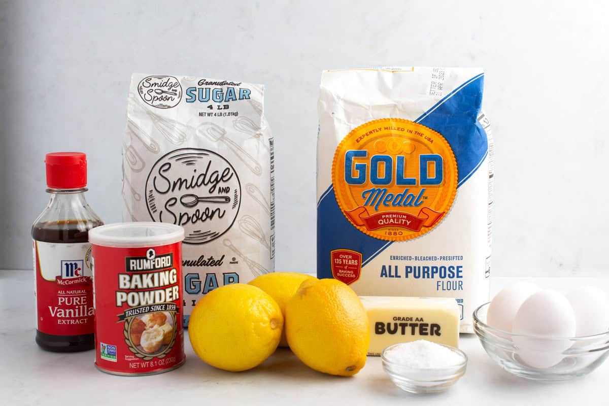 Flour, sugar, vanilla extract, baking powder, three lemons, a stick of butter, a small bowl of salt, and three eggs on a white countertop.