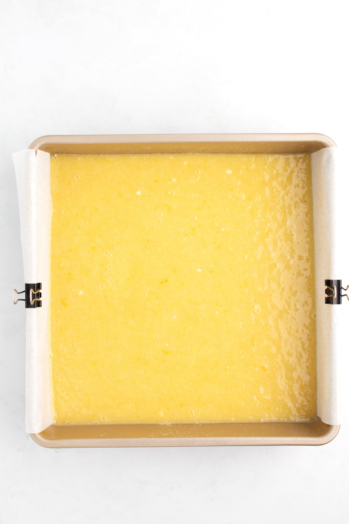 A square cake pan with parchment paper sling and lemonie batter inside.