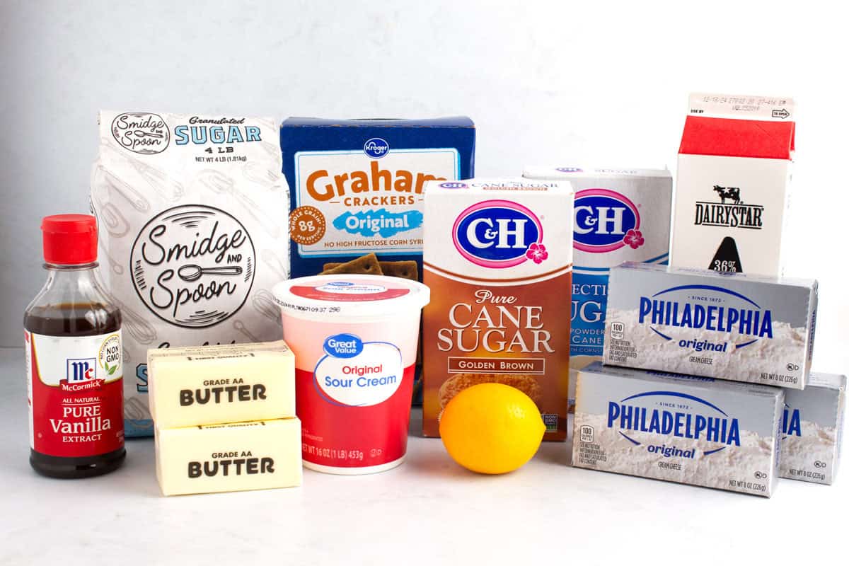 Powdered, granulated, and brown sugar, graham crackers, heavy cream, cream cheese, lemon, sour cream, butter, and vanilla on counter.
