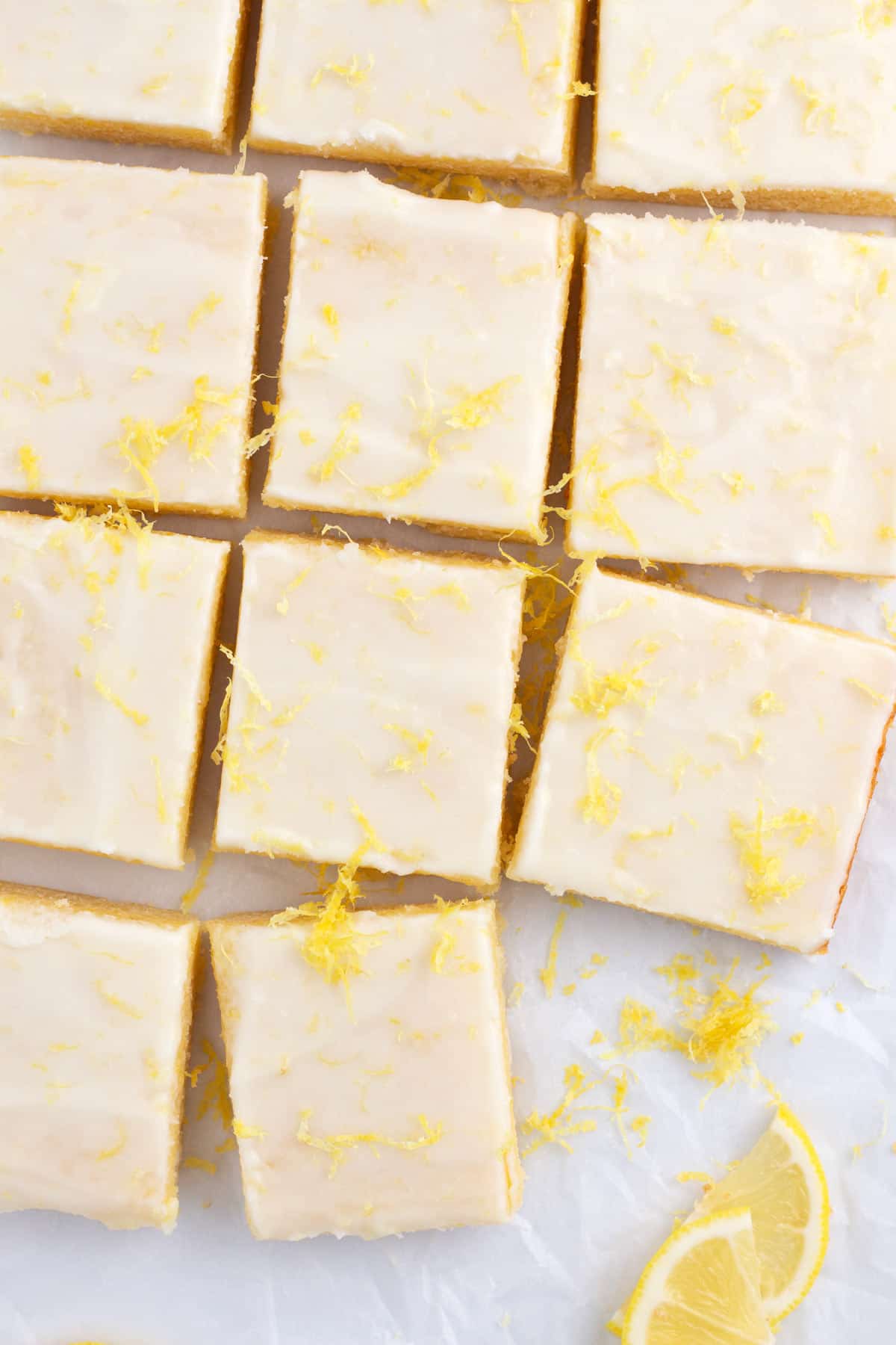 Rows of lemonies with lemon frosting randomly spaced on parchment paper with lemon zest and slices around.
