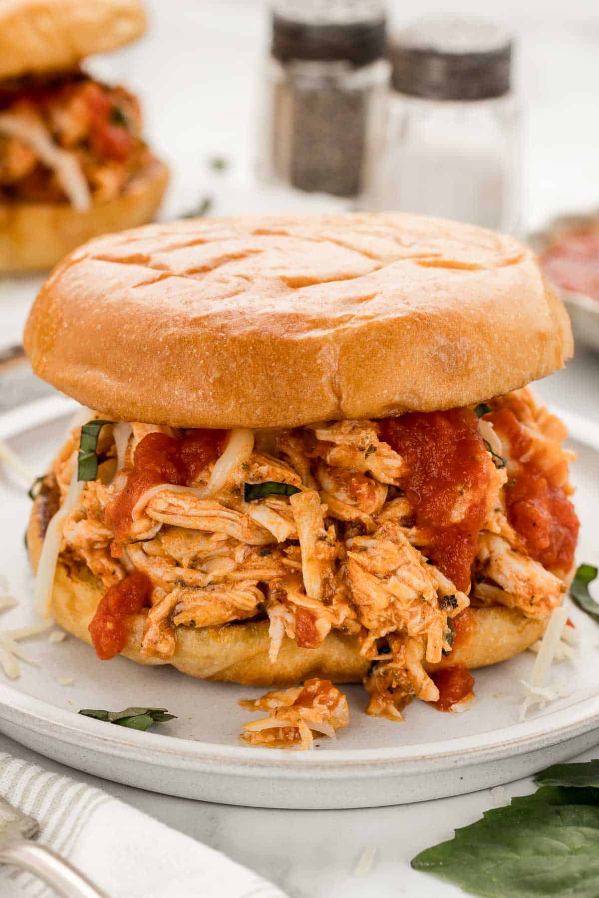 A marinara chicken sandwich on a plate with a small bowl of sauce behind it and a towel with a fork in the bottom left corner.