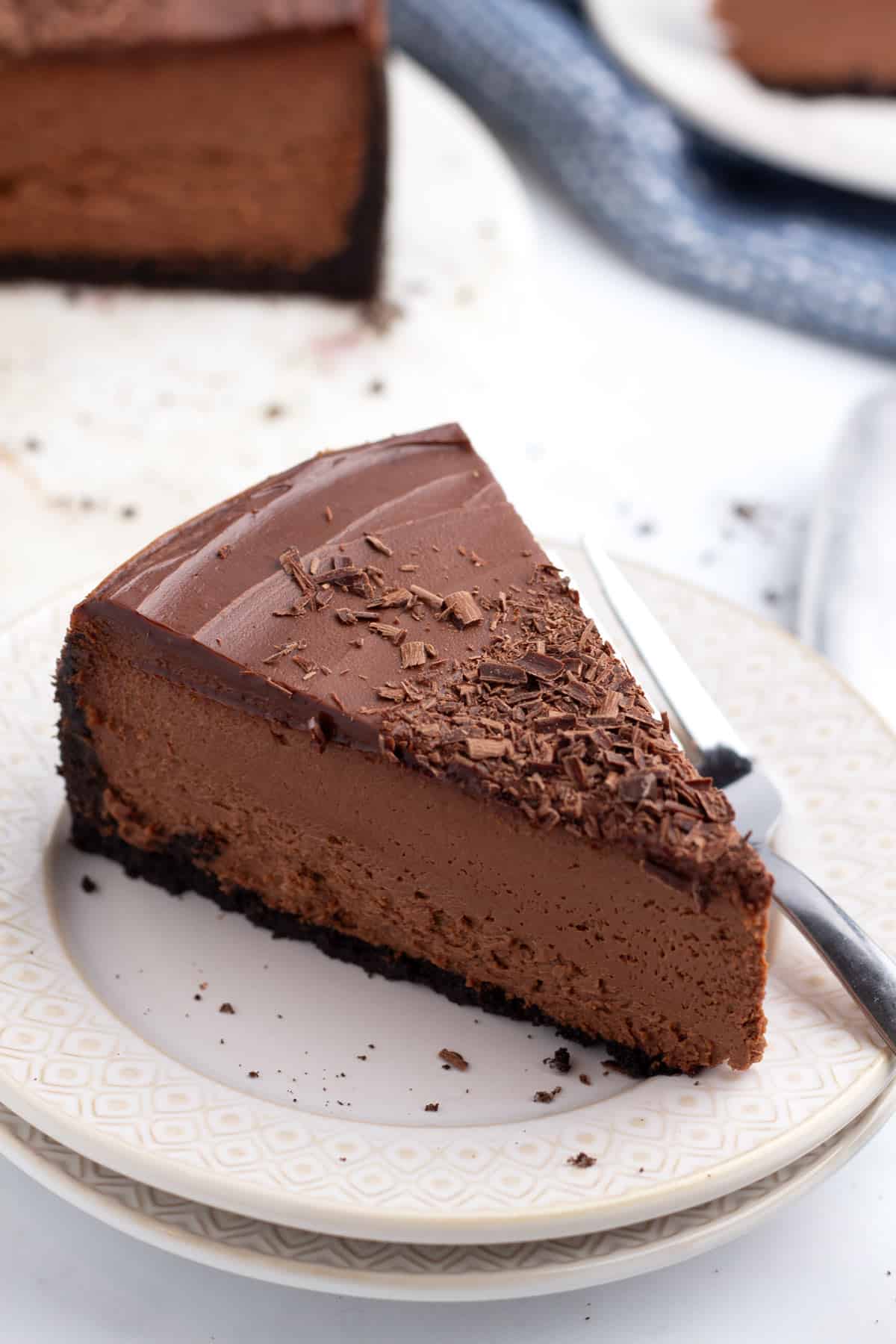 Slice of a chocolate cheesecake on a stack of small plates with a fork next to it and two more slices of cheesecake on plates with a blue towel in the background.