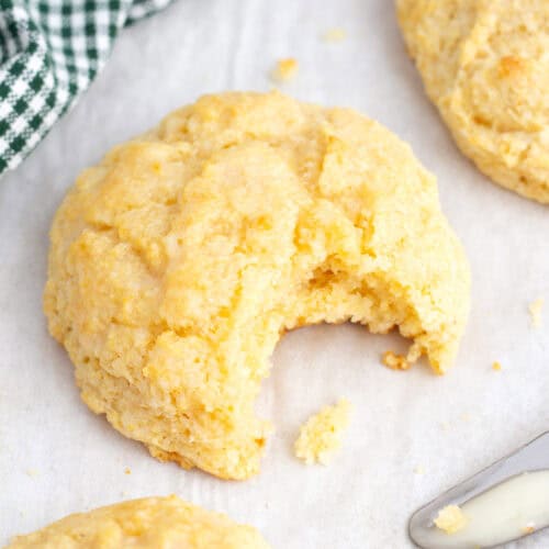 A very close up view of a yellow, textured biscuit with a bite removed on white parchment paper with the edge of a silver butter knife in the front of the photo.