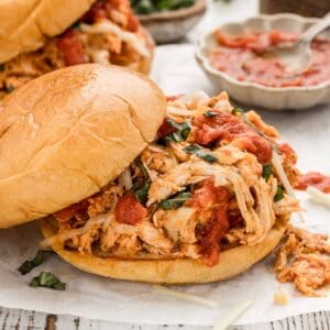 Shredded marinara chicken with the top bun sliding off with another sandwich next to it and a bowl of sauce and a bowl of herbs behind it.