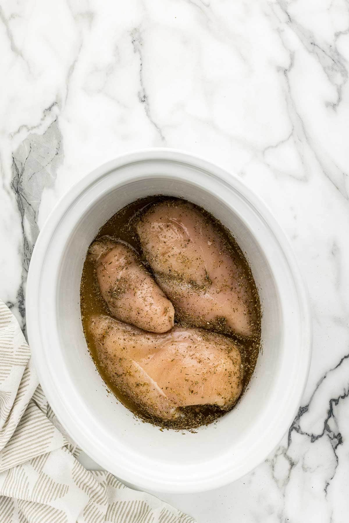 A white slow cooker pot with chicken breasts and broth in it on a marble counter with a white towel in the bottom left corner.