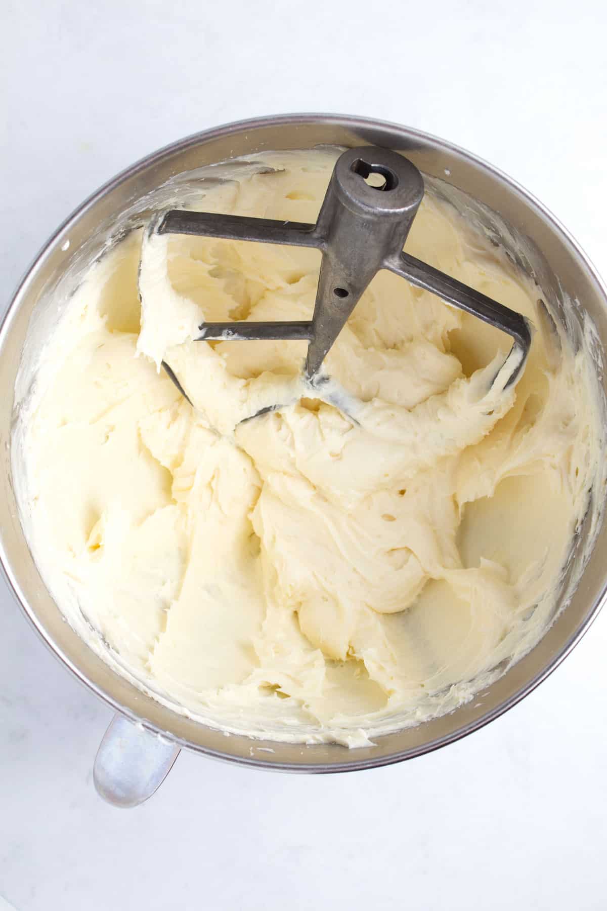 White batter in a stainless steel bowl being blended with a stand mixer.