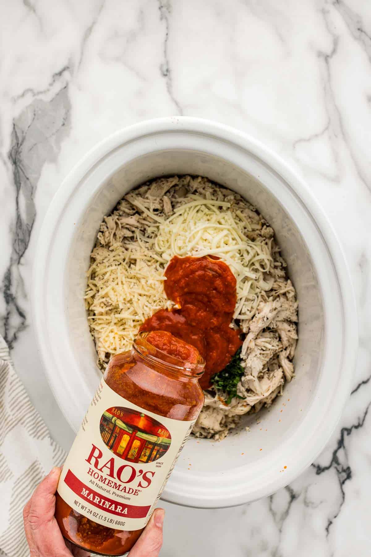 Shredded chicken, cheese, and herbs in a white slow cooker pot on a marble counter with a jar of marinara sauce being poured into the pot and a striped towel in the bottom left corner.
