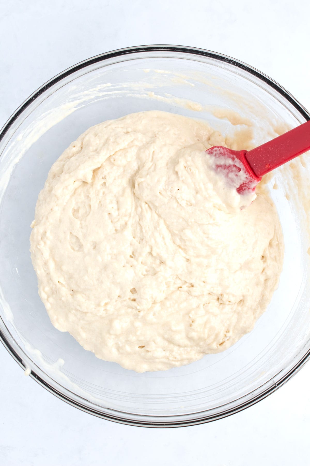 A large clear glass bowl with a wet bread dough and a red spatula resting on the side.