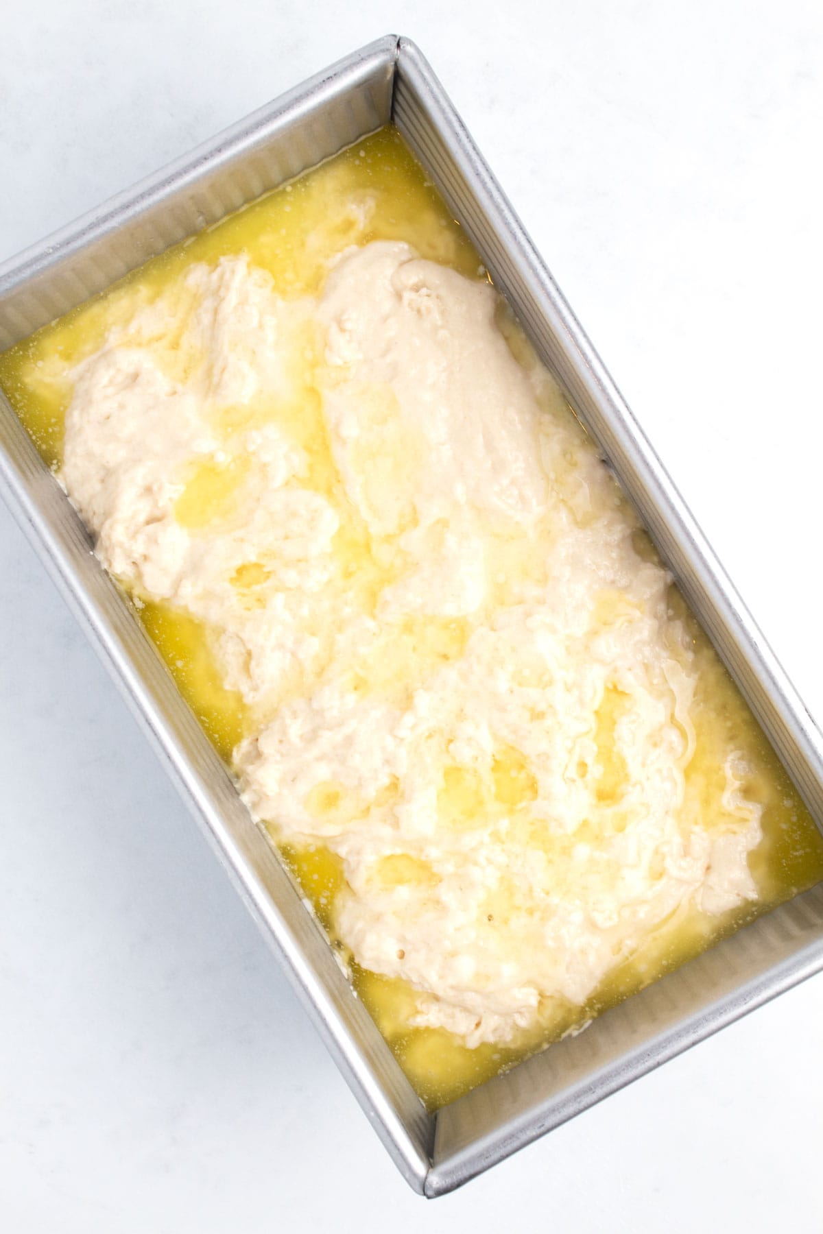 A silver loaf pan half full of a white batter with melted butter poured on the top.