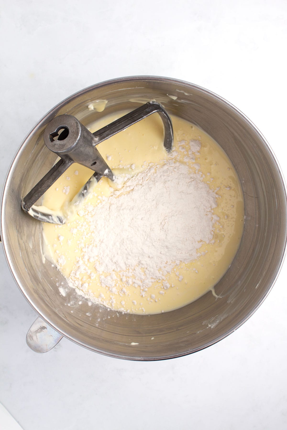 Stainless steel bowl with batter and unmixed powder and a stand mixer paddle in the top left part of the bowl.