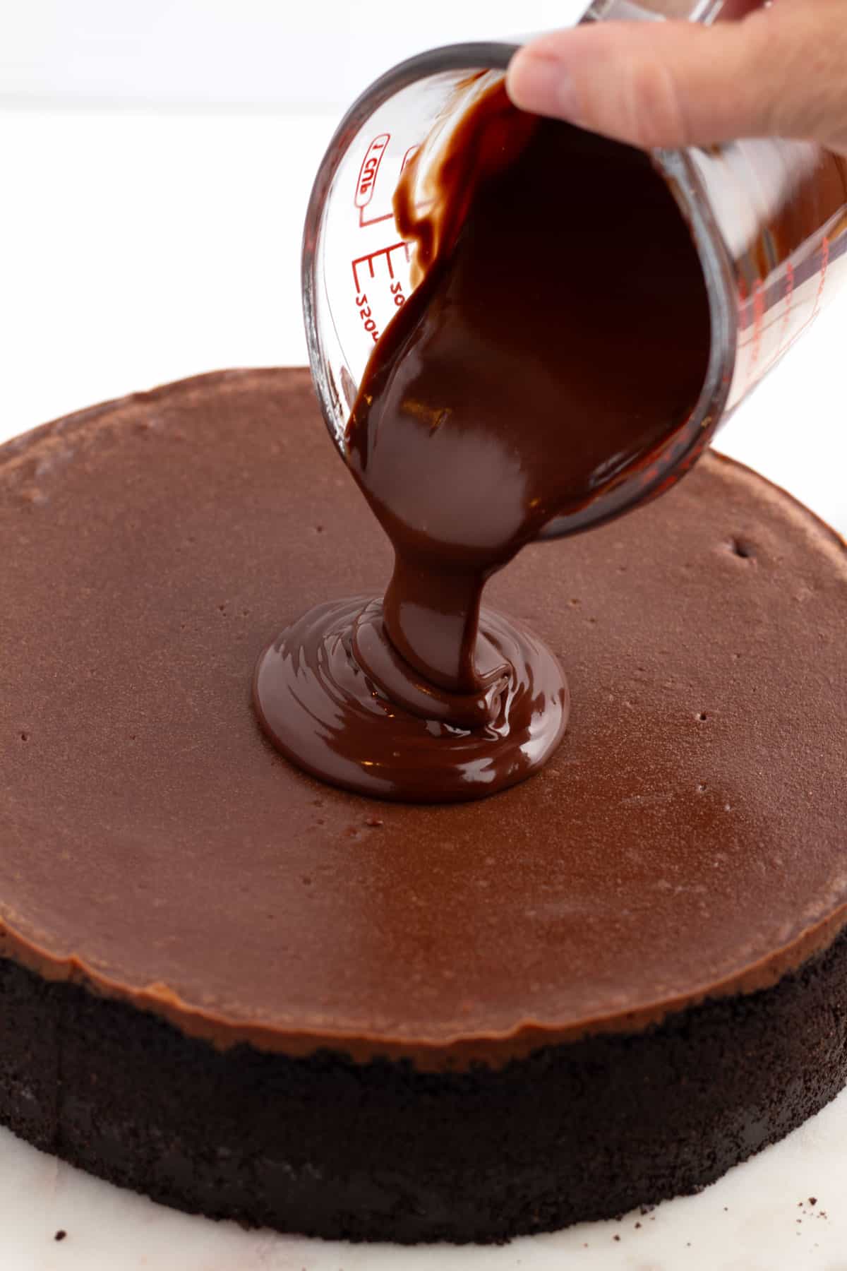 Bare chocolate cheesecake with chocolate frosting getting poured onto it from a small glass measuring cup.