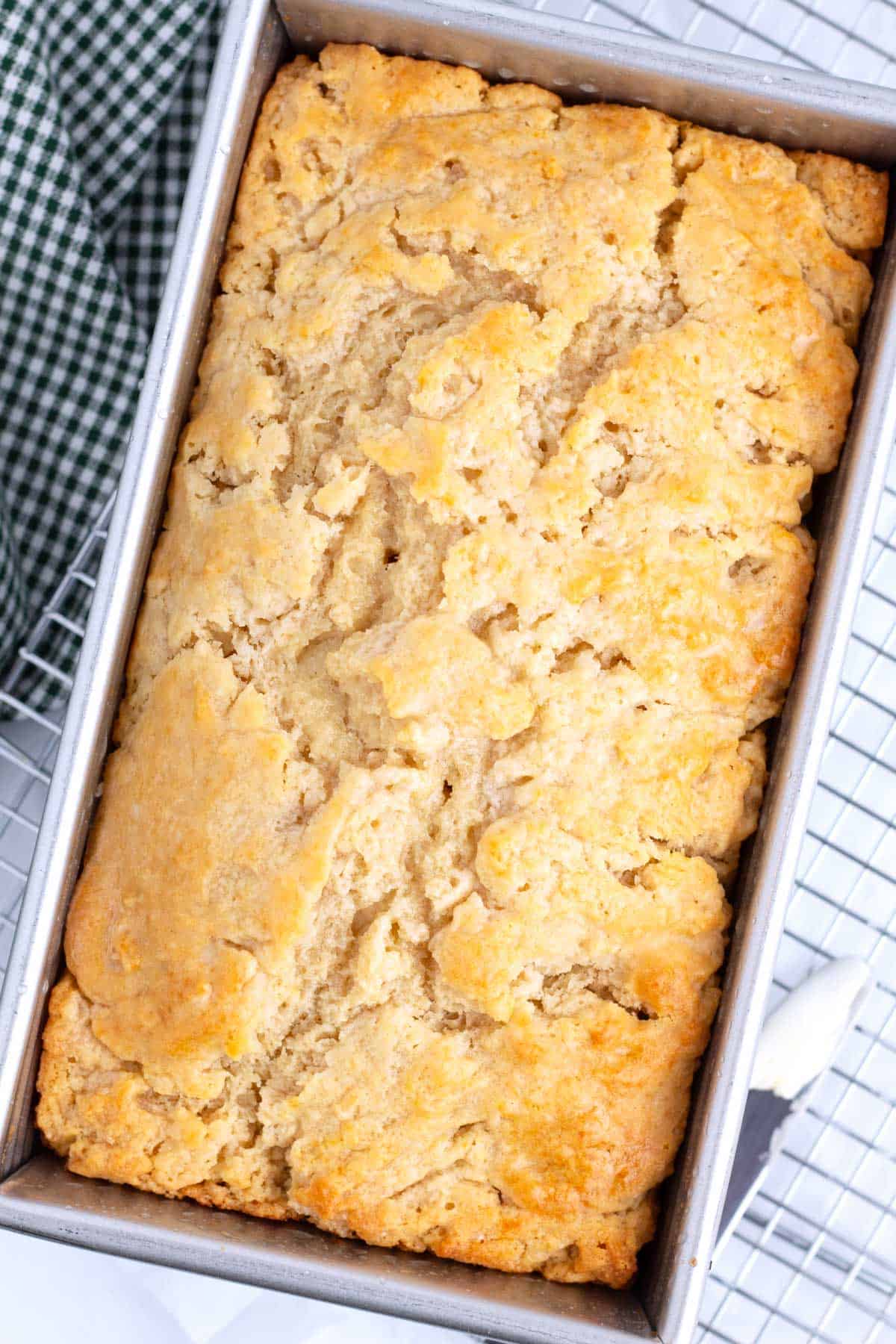 Golden brown, crinkled bread in a silver loaf pan sitting on top of a wire rack at a diagonal angle with a green checkered towel on the left side of the pan.