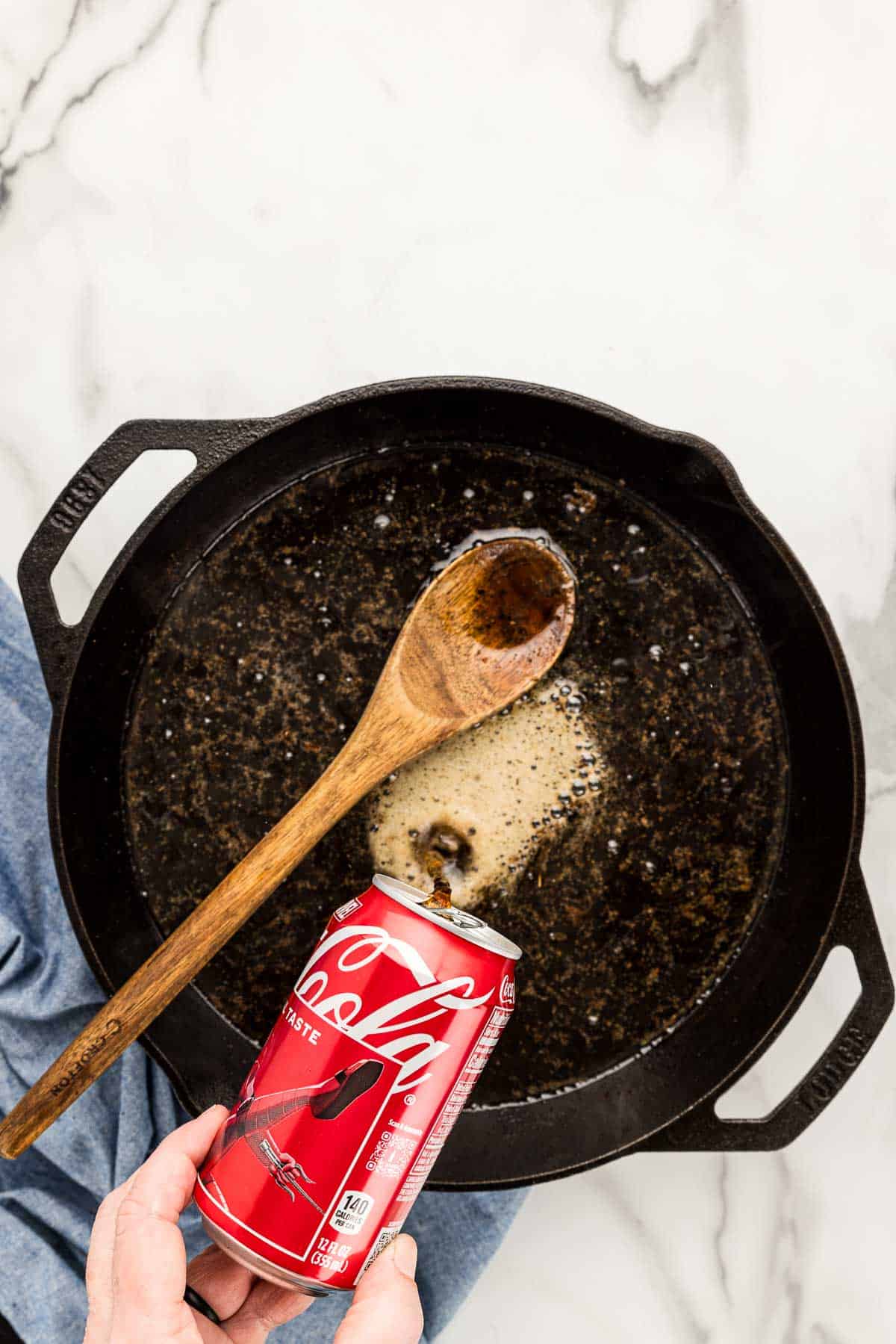 A can of Coca Cola being poured into a cast iron skillet with a wooden spoon.