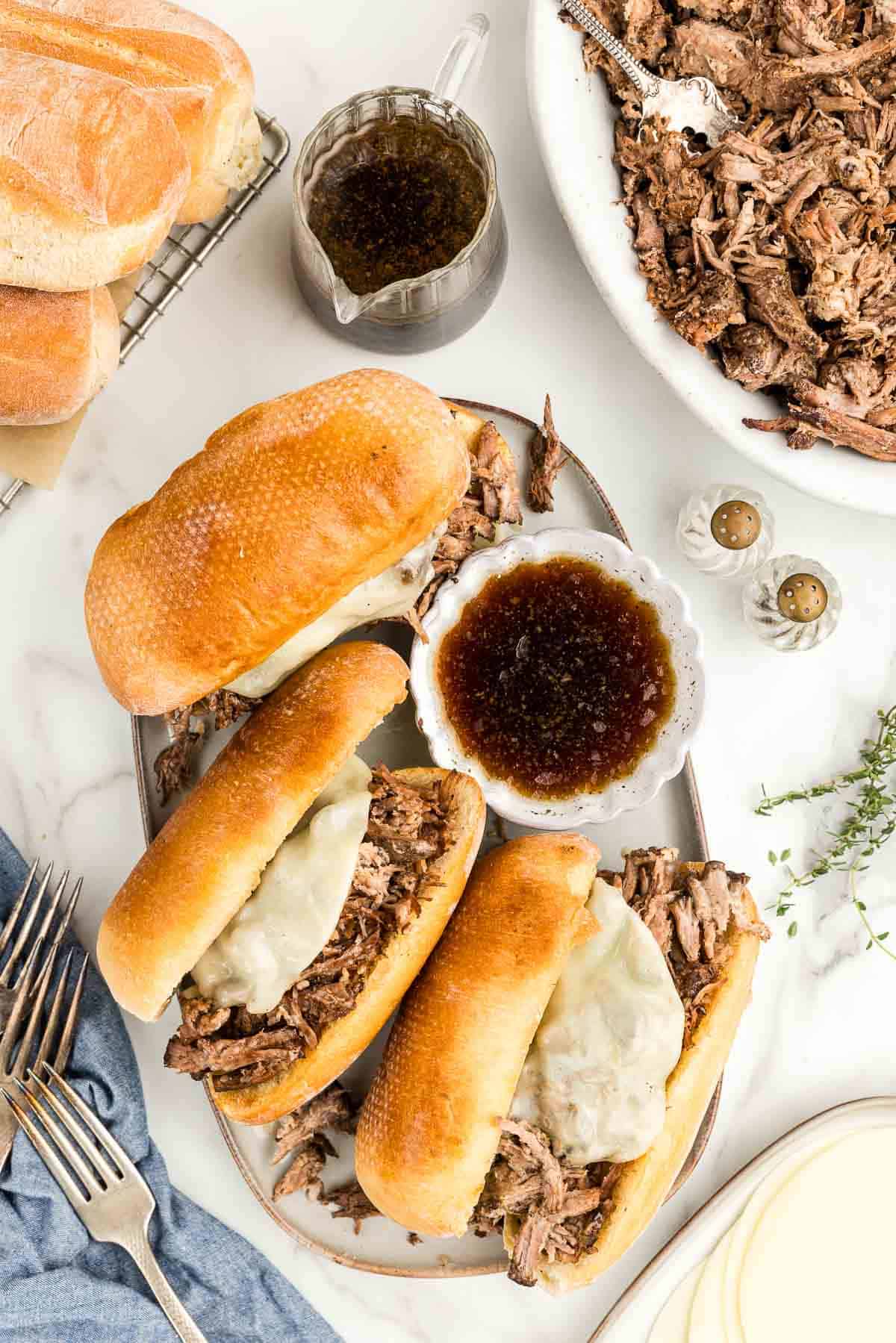 A tray of three chuck roast sandwiches and a bowl of French dip surrounded by toppings and utensils.