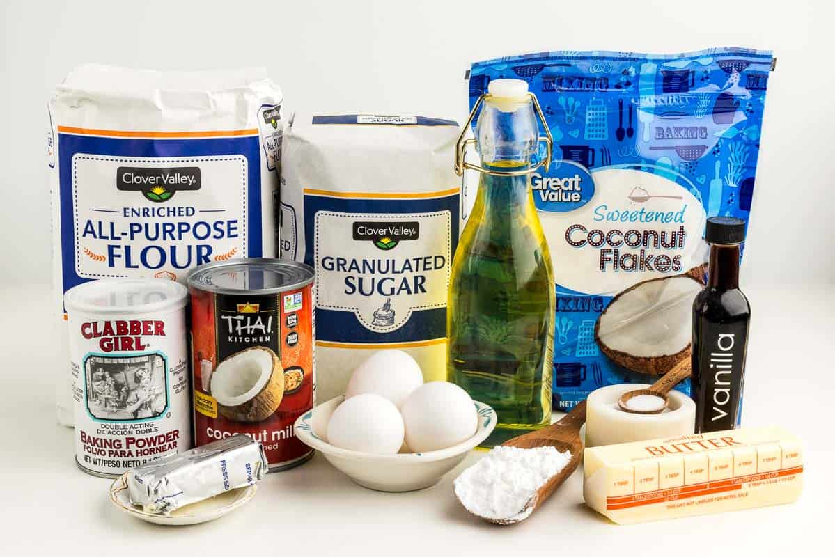 Baking powder, flour, coconut milk, cream cheese, sugar, eggs, oil, a stick of butter, coconut flakes, vanilla, powdered sugar, and salt on a white surface.