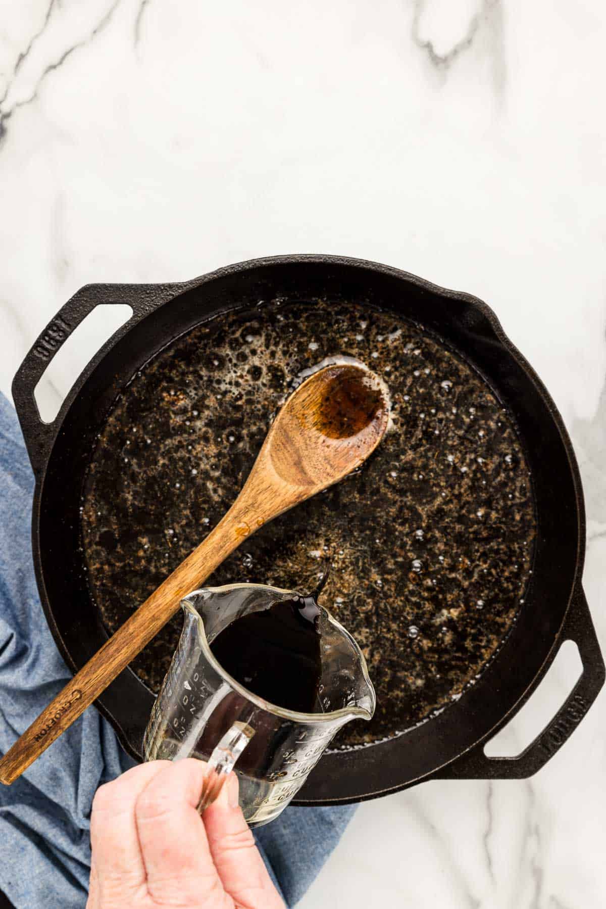 A skillet with a measuring cup of sauce being poured and a cooking spoon.