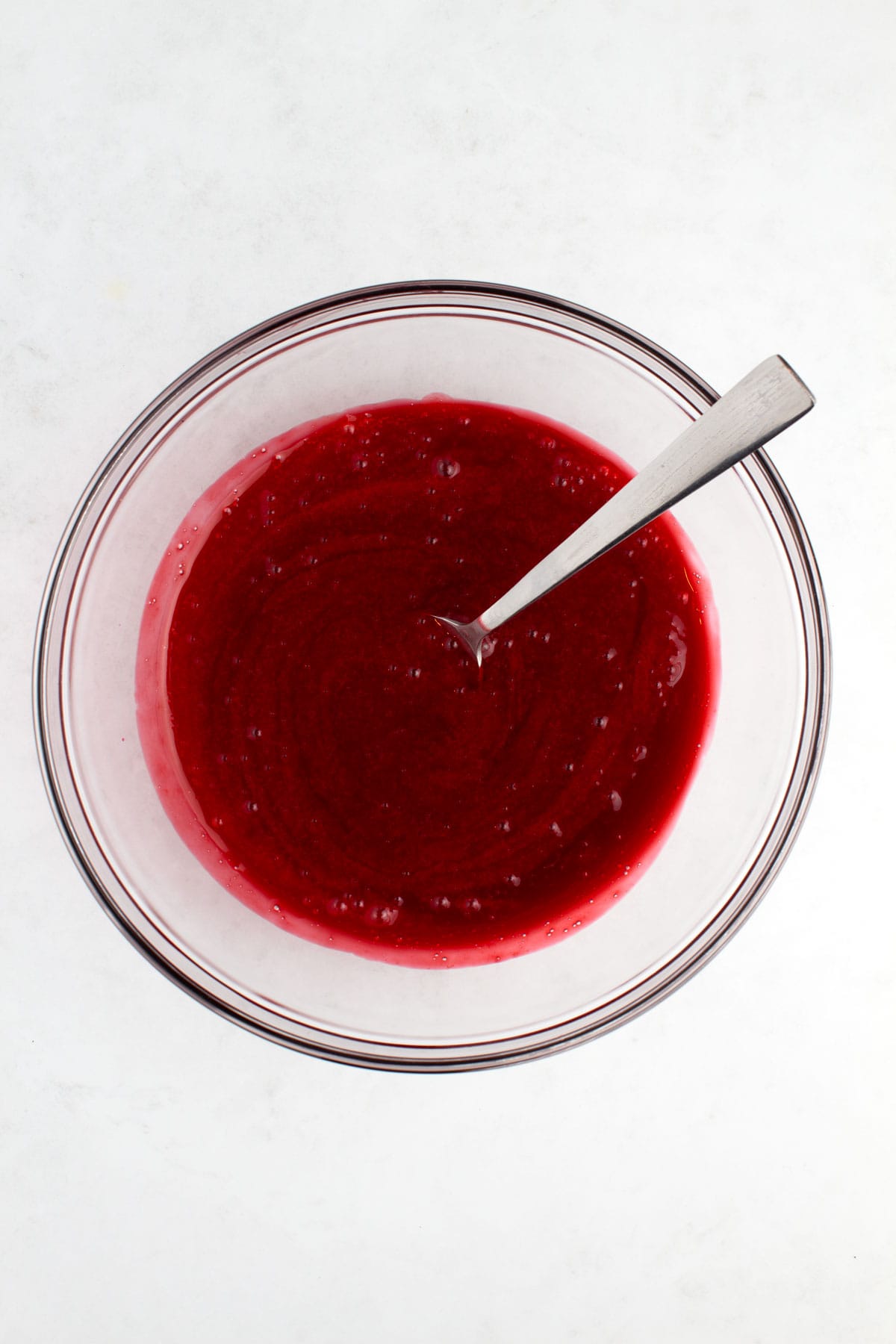 A glass bowl of raspberry sauce with a spoon resting in it.