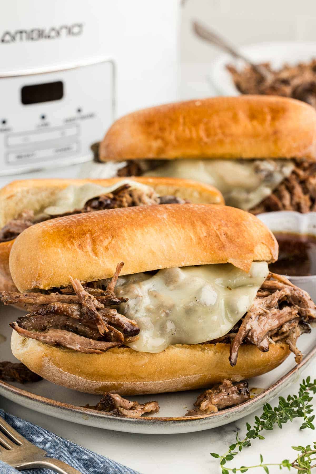 Three juicy French dip sandwiches on a tray in front of a slow cooker.