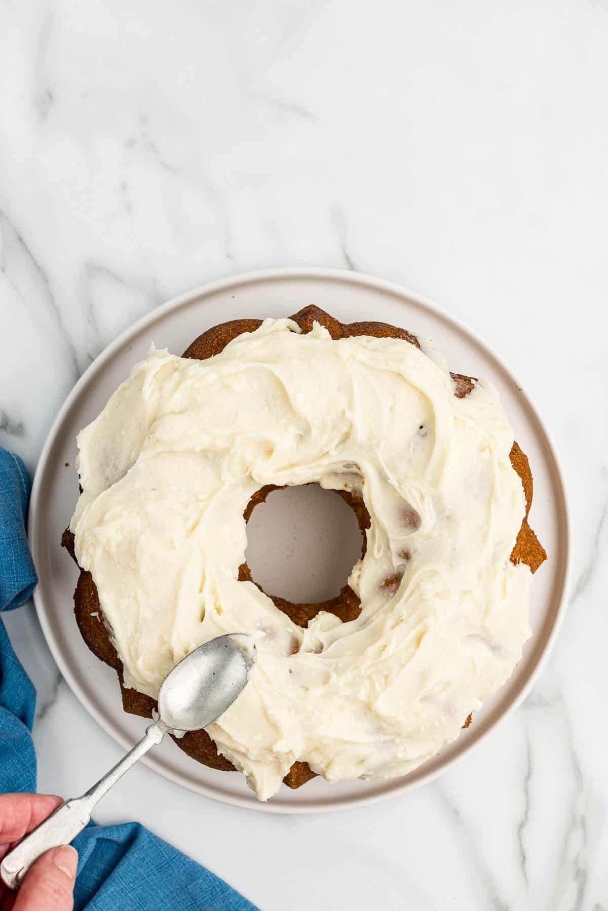 Bundt cake with white frosting on top getting smoothed out with a spoon.