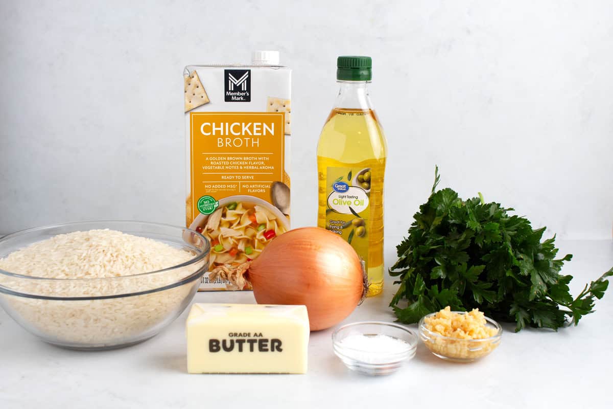 A bowl of rice, a stick of butter, an onion, a dish of salt, garlic, herbs, olive oil, and chicken broth on a white surface.