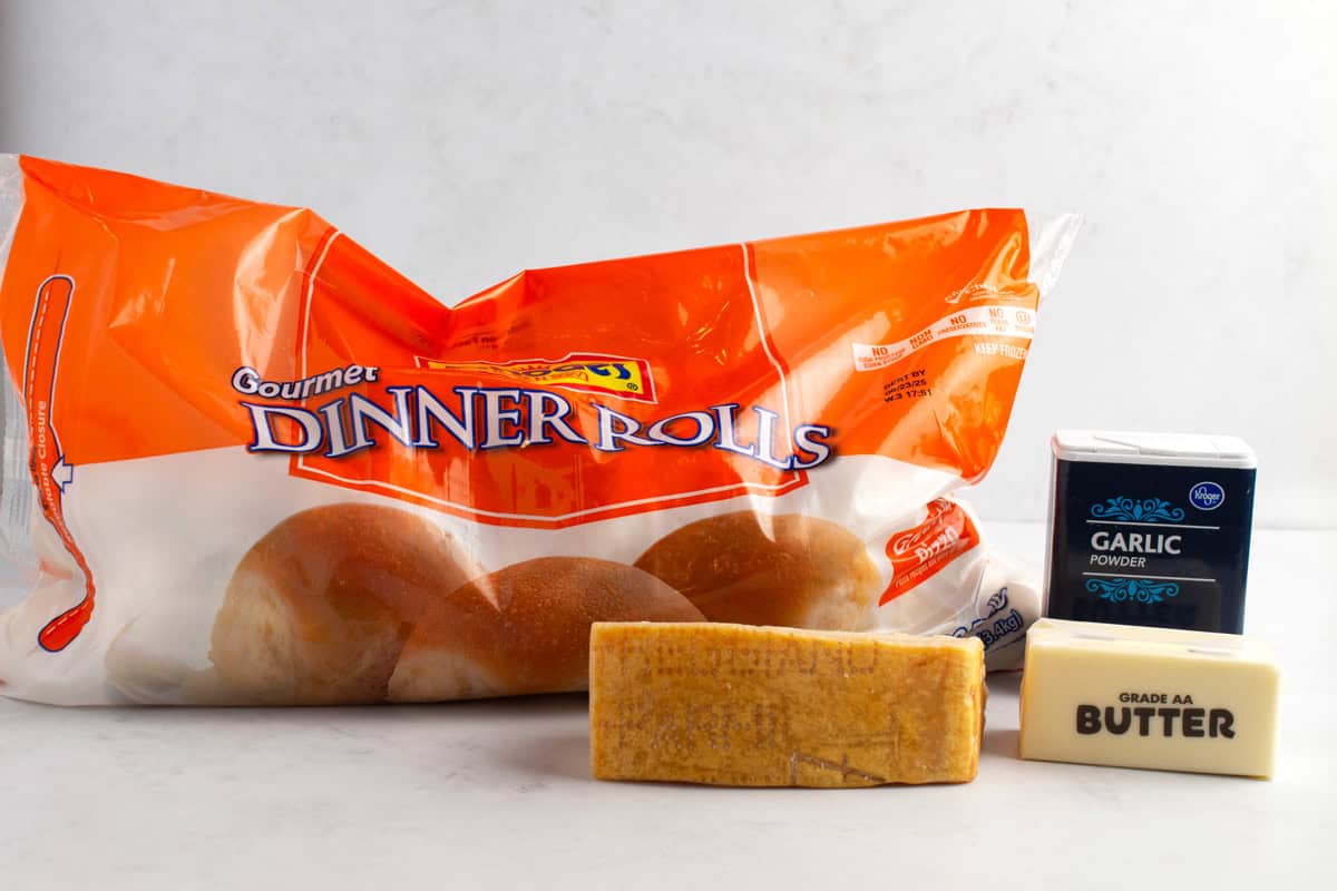 Frozen dinner rolls, a stick of butter, garlic powder, and parmesan cheese on a white counter.