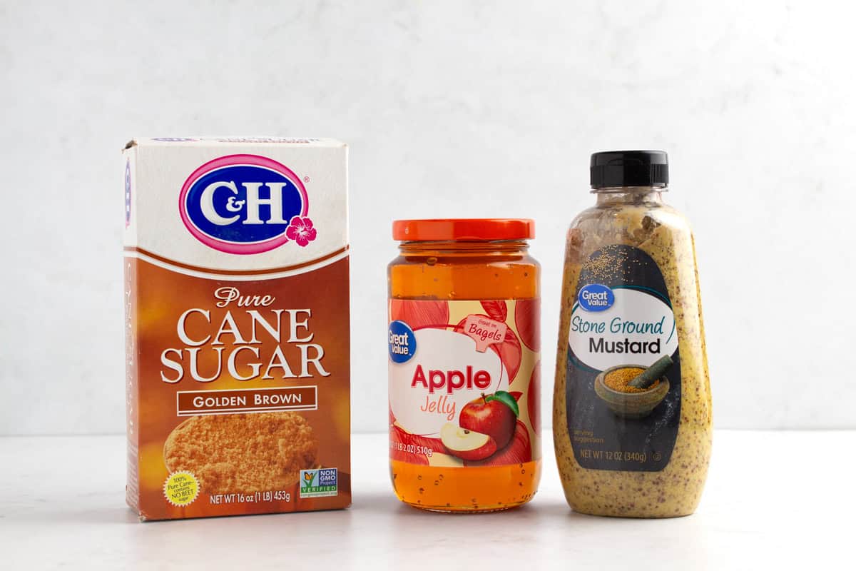 Cane sugar, a jar of apple jelly, and a bottle of ground mustard on a white counter.