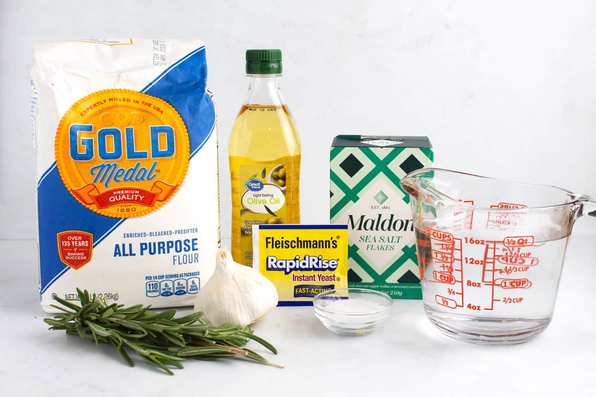 Flour, rosemary, garlic, olive oil, yeast, a small bowl of salt, sea salt flakes, and two cups of water in a measuring glass on a white counter.