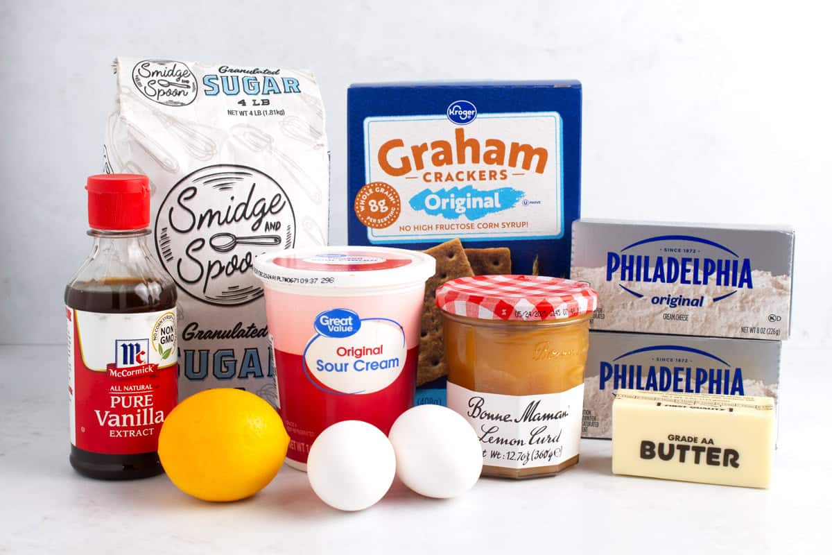 Vanilla extract, sugar, a lemon, two eggs, sour cream, a box of graham crackers, a jar of lemon curd, a stick of butter, and two boxes of cream cheese on a counter.