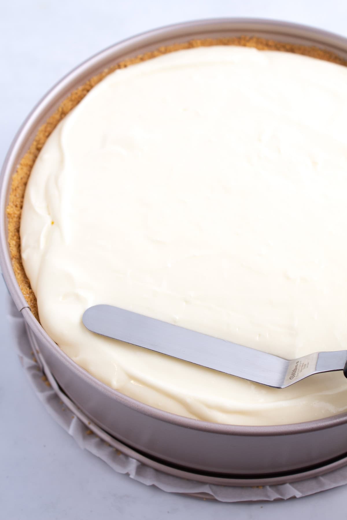 A steel spatula smoothing cheesecake batter into a circular baking dish.