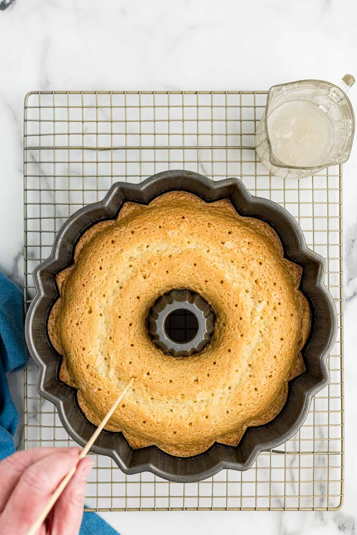 Bundt cake in a baking pan with holes getting poked in the top with a toothpick and a small glass of coconut syrup next to it.