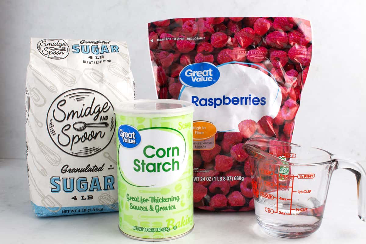 Sugar, corn starch, frozen raspberries, and a measuring glass of water on a white counter.