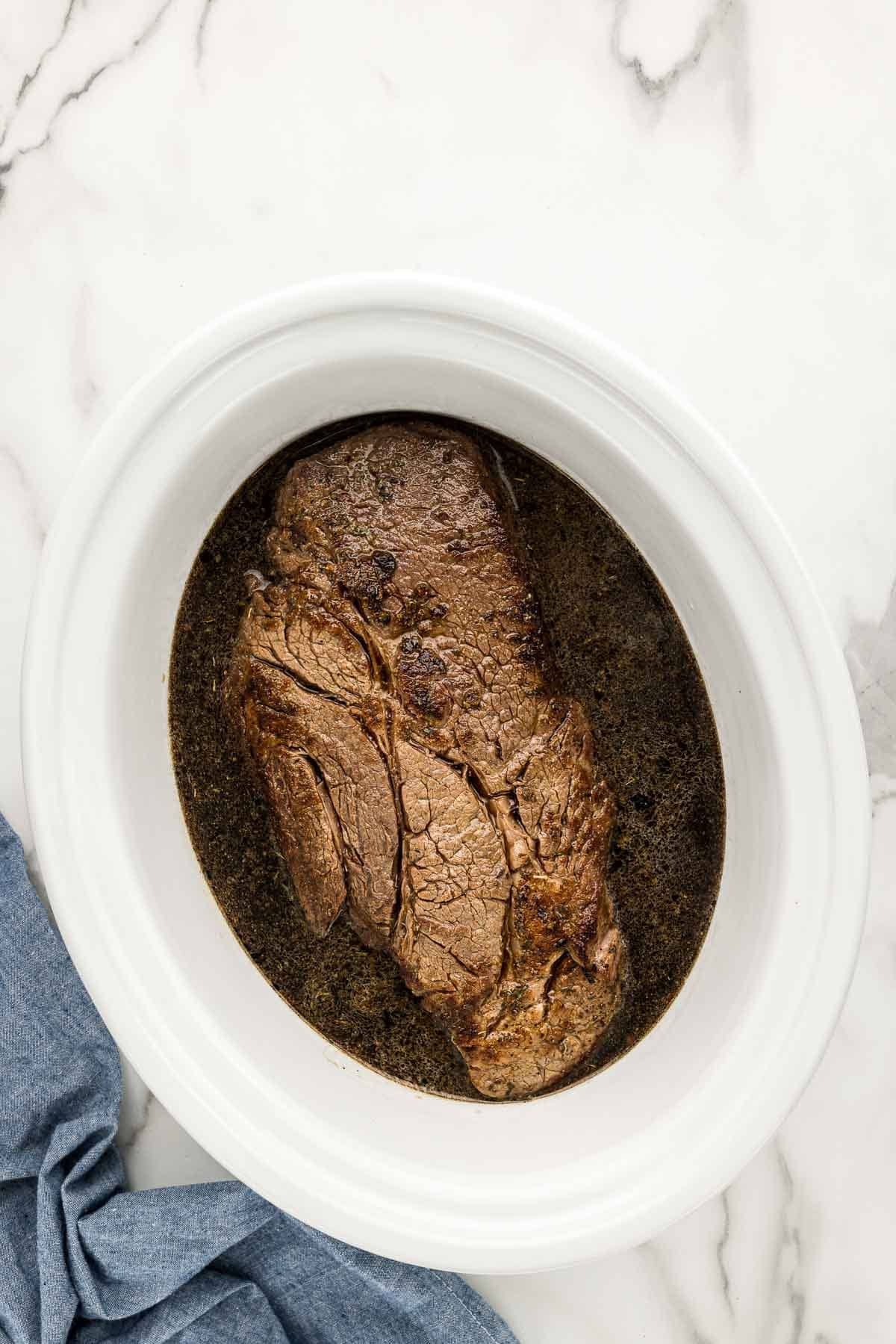A large chuck roast sitting in a crockpot and floating in juices.