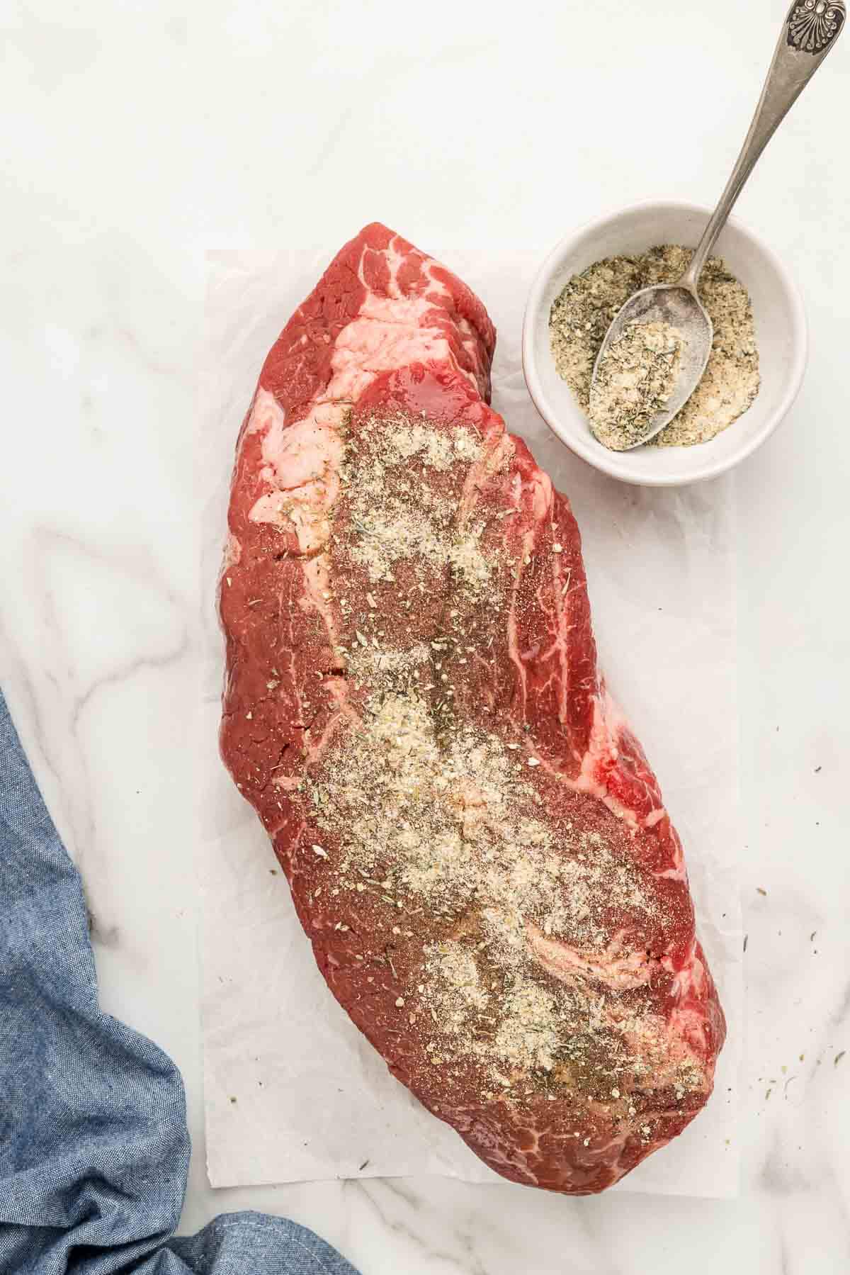 A piece of raw meat sprinkled with seasonings with a bowl and spoon next to it.