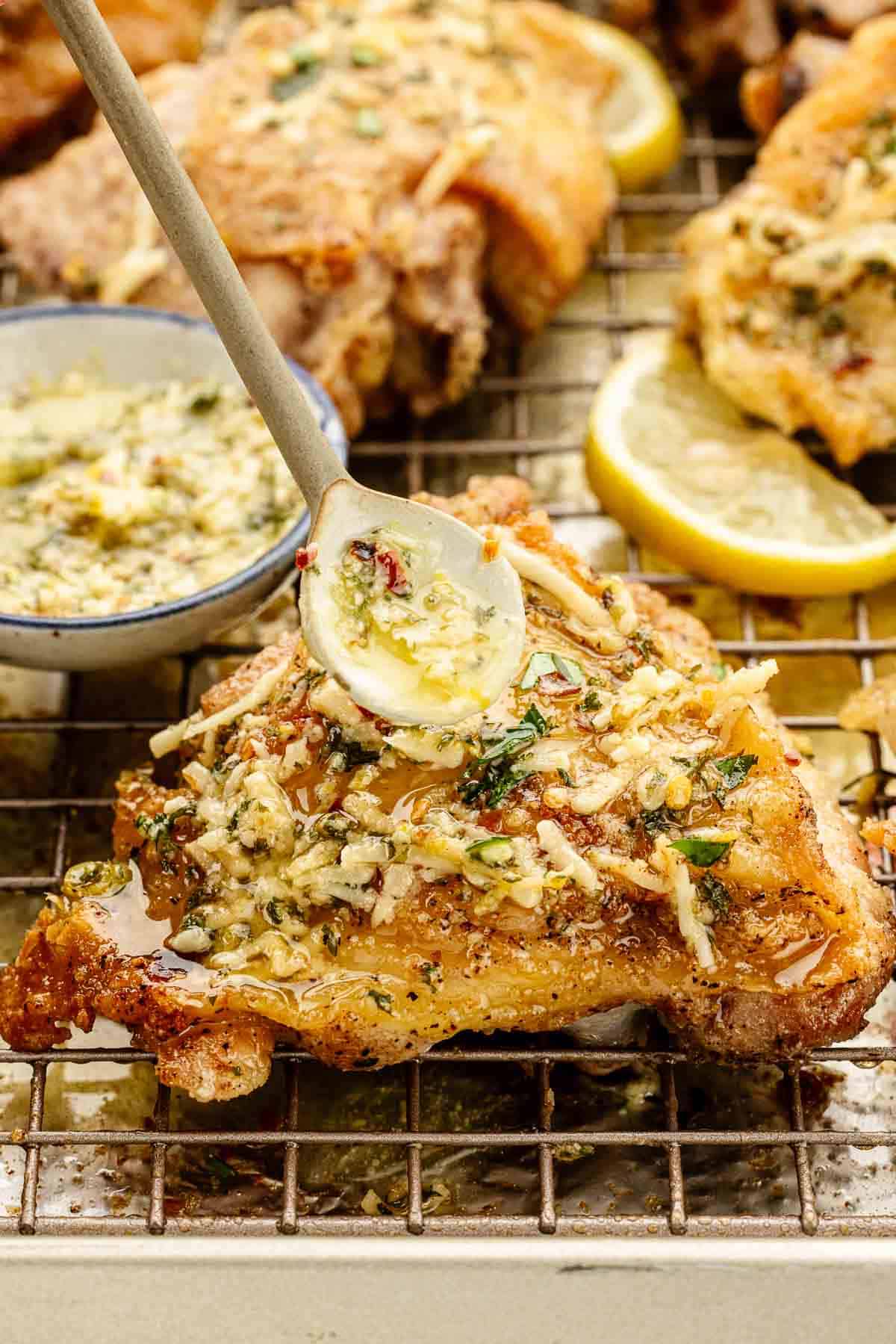 A baked chicken thigh on a cooling rack with sauce dripping on it from a small spoon.
