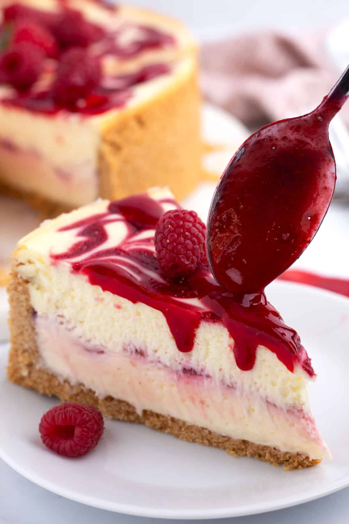A piece of cheesecake on a white plate with a spoon drizzling raspberry topping on it.