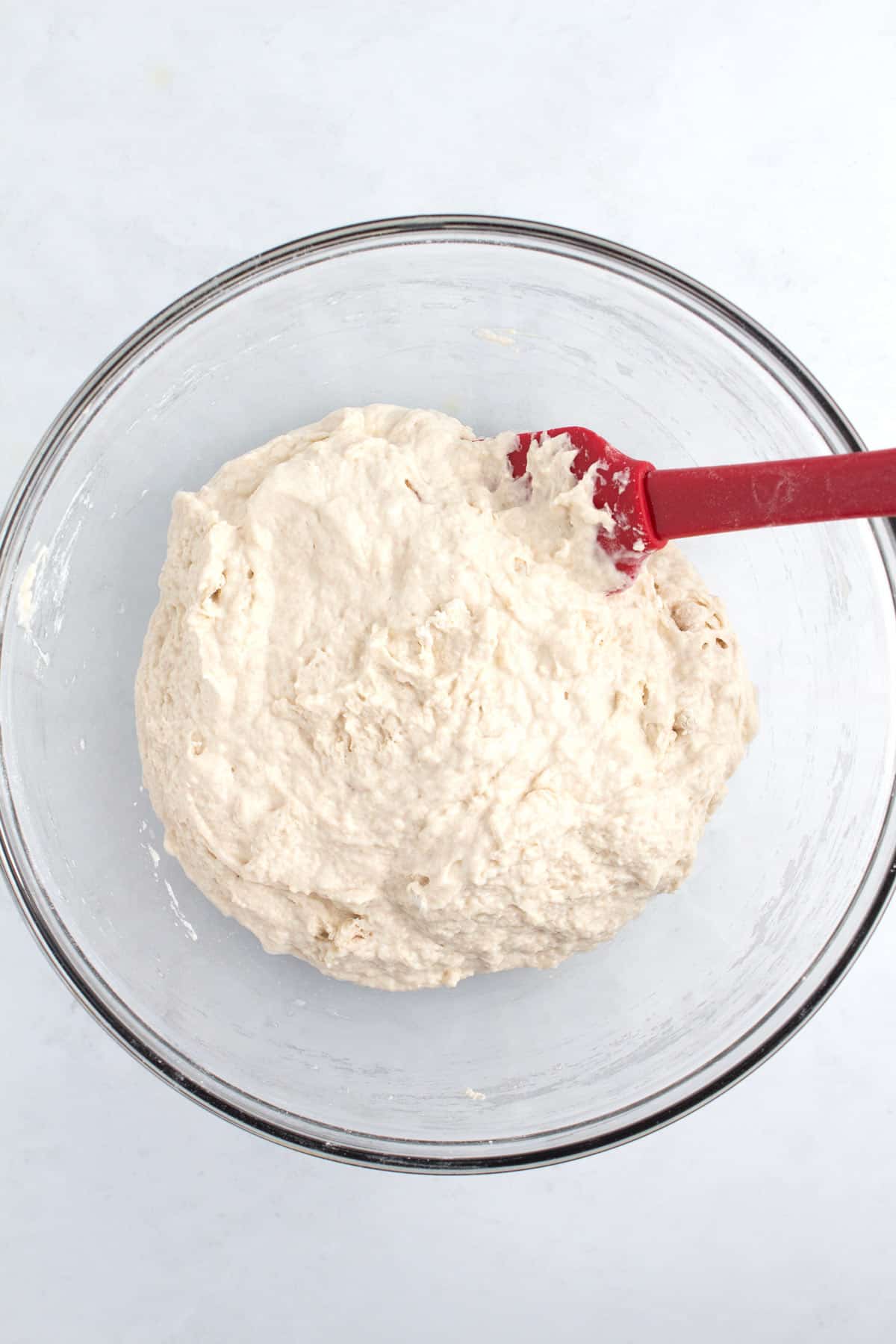A glass bowl of dough with a red spatula stuck in the upper corner.