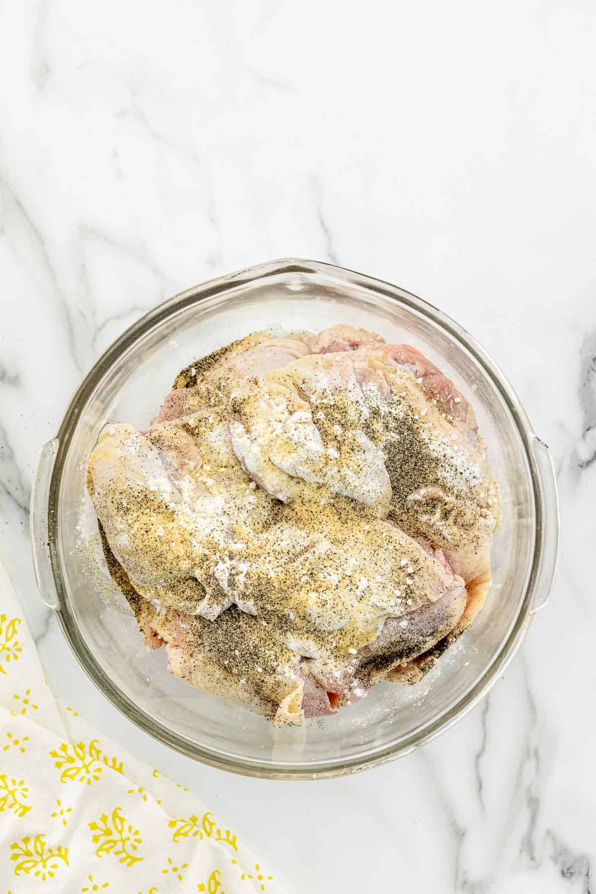 A mixing bowl full of raw chicken coated with seasoning on a marble counter.