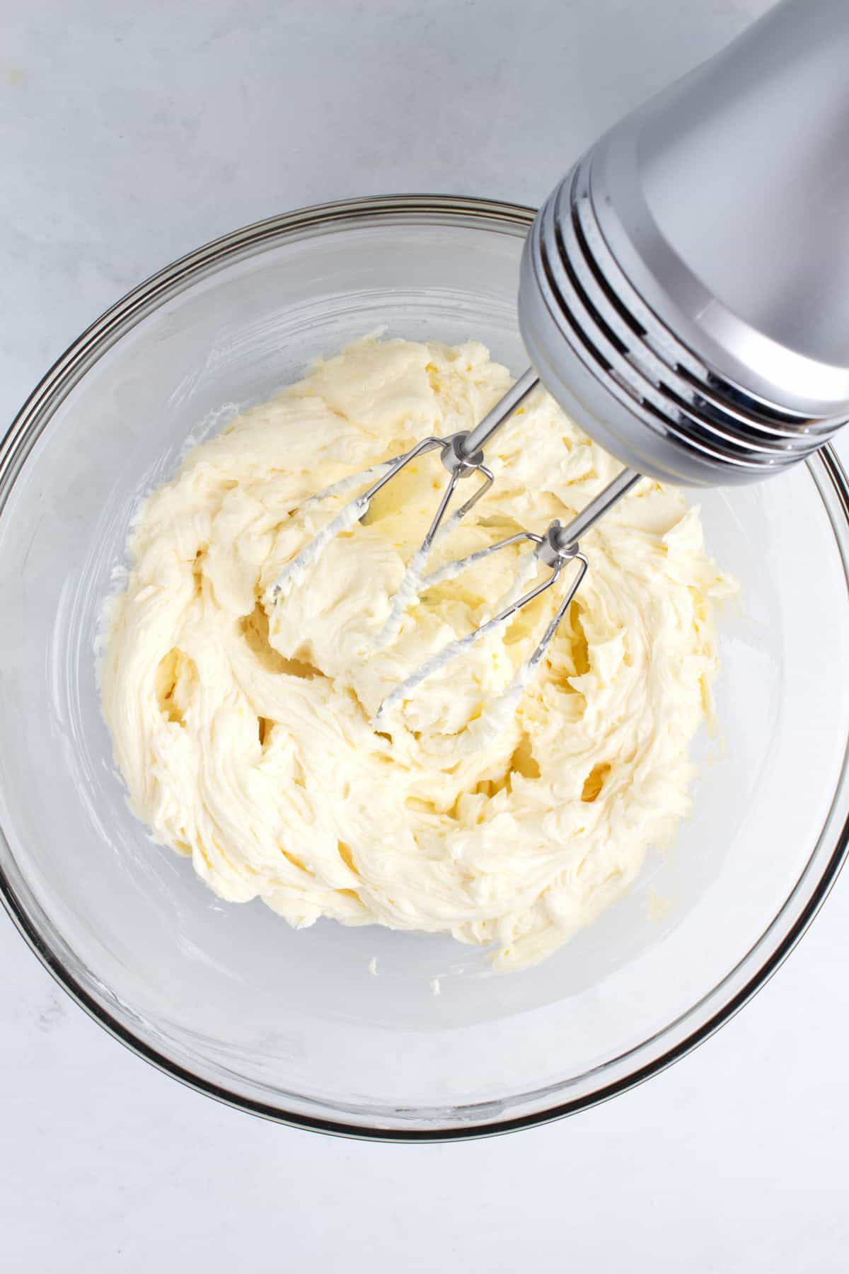 A glass bowl of cream cheese batter with a silver hand blender.