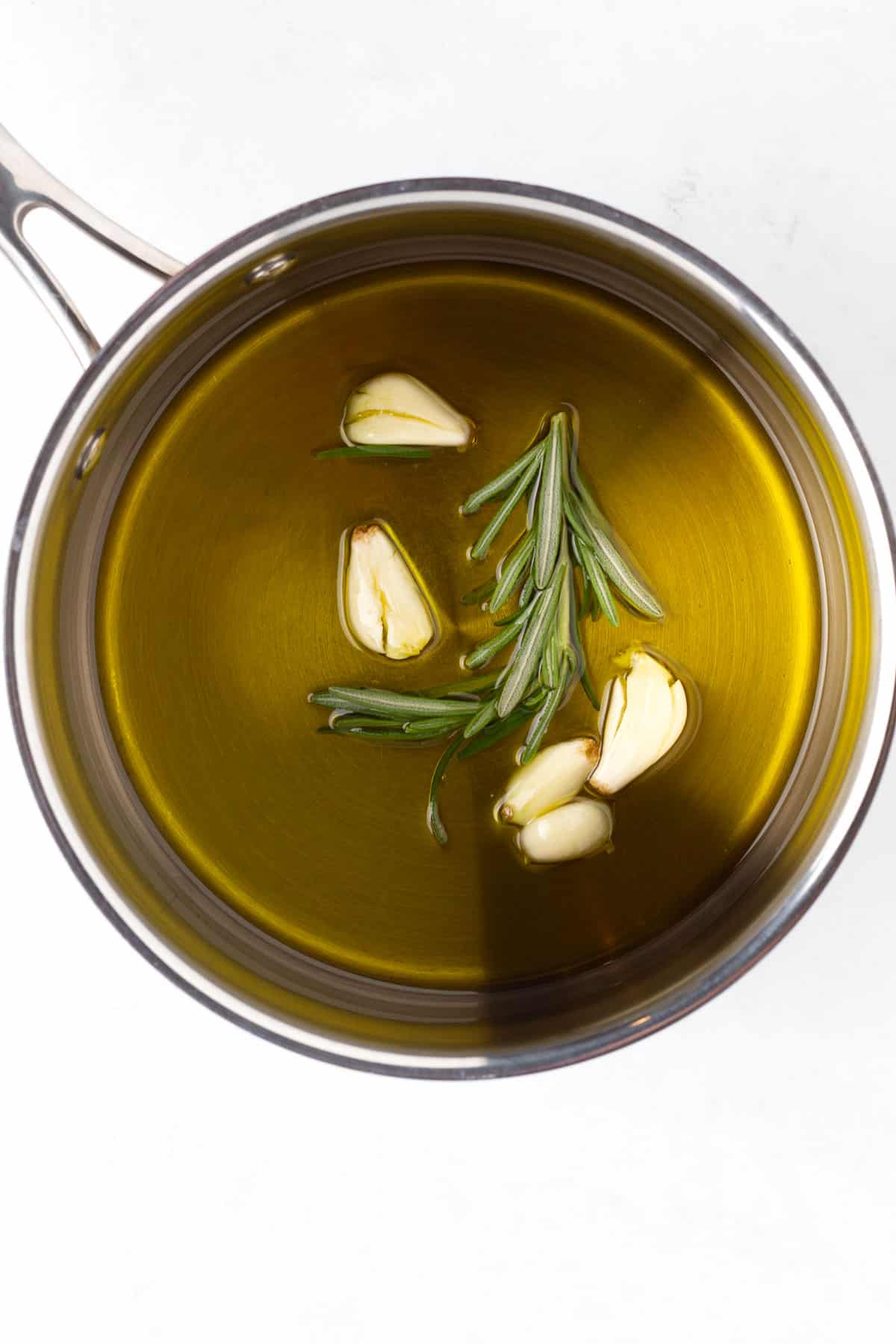 A saucepan full of olive oil with rosemary and garlic floating in it.