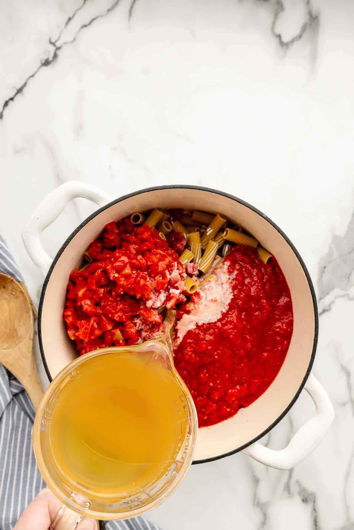 A pot full of sauce and noodles with a measuring cup pouring broth into it.
