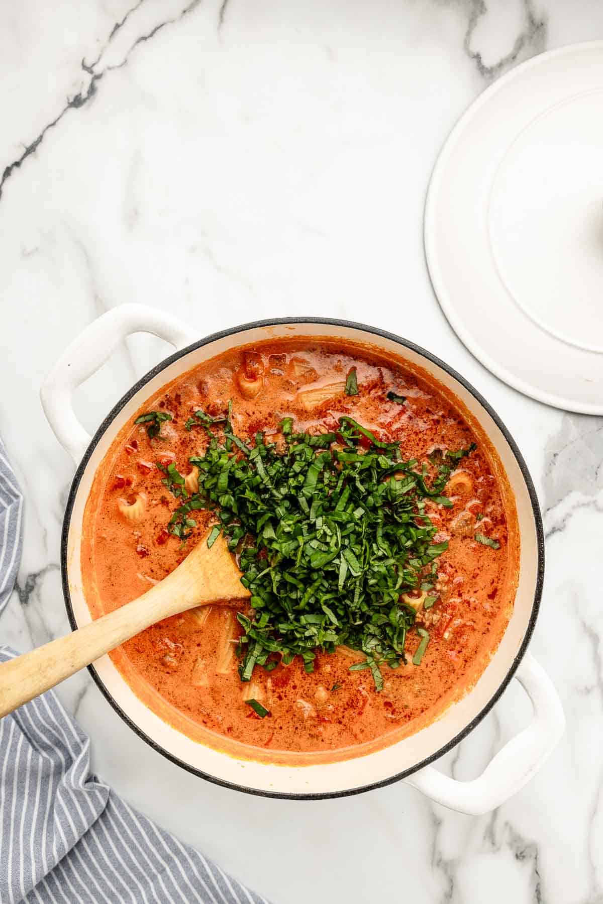 Mixed pasta and sauce in a pot with herbs and a wooden spoon resting in it.