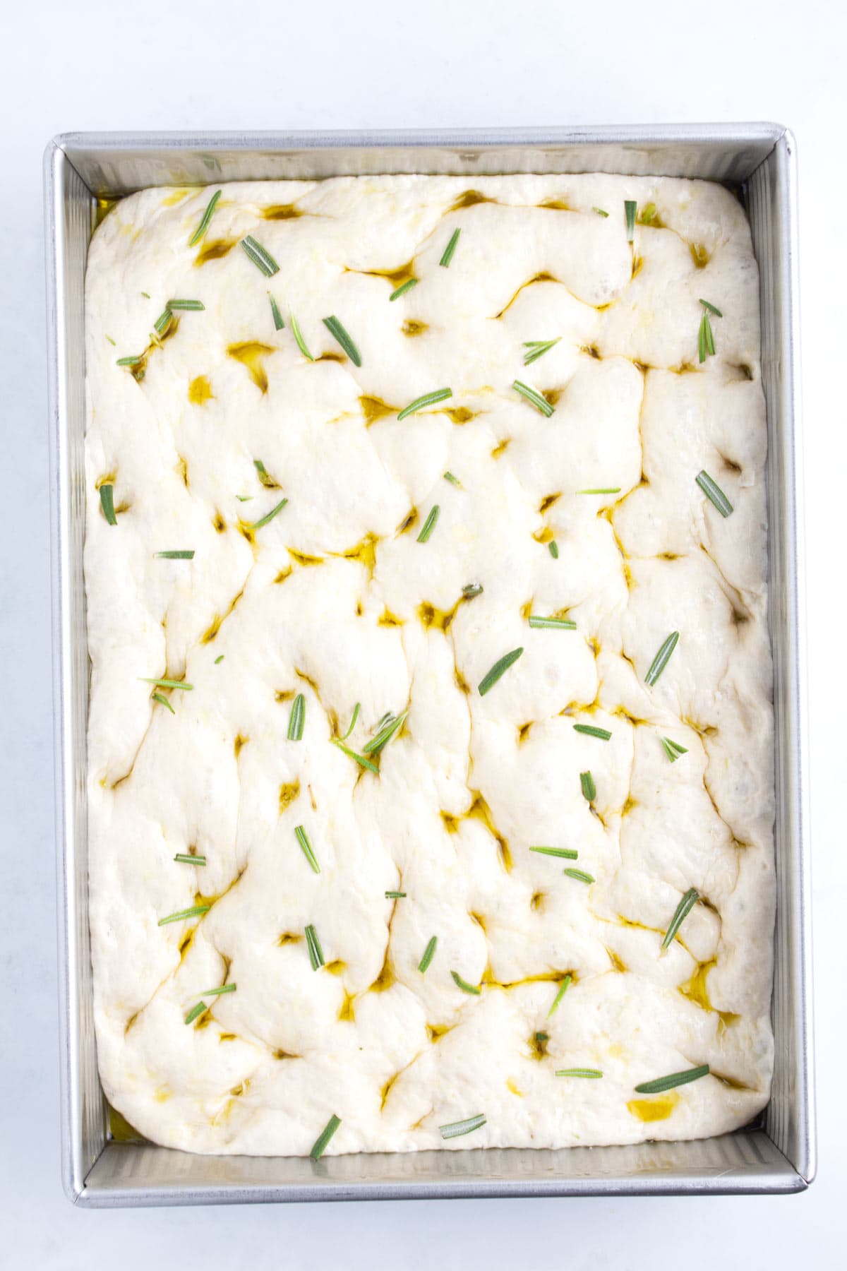 Raw bread dough in a rectangular pan sprinkled with rosemary and olive oil.