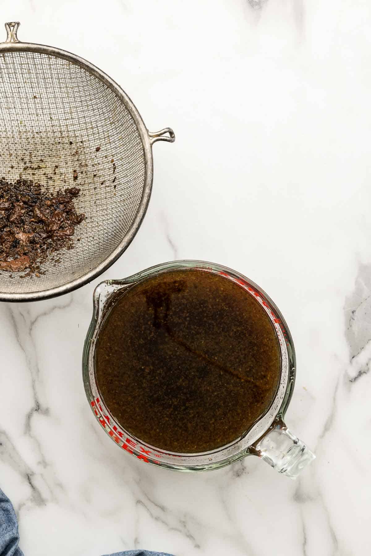 A small glass measuring cup of dip with a wire mesh strainer beside it.