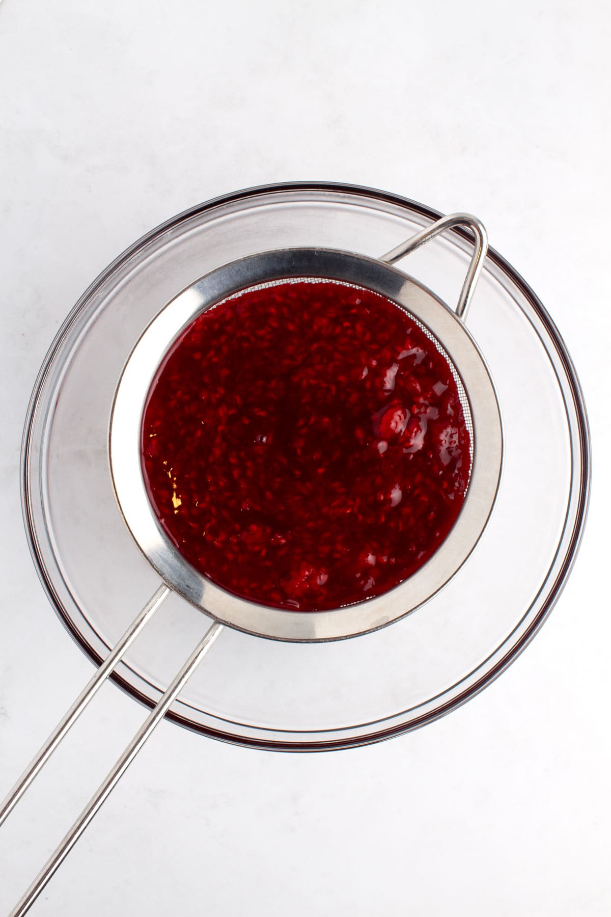 Raspberry topping getting strained over a glass mixing bowl.