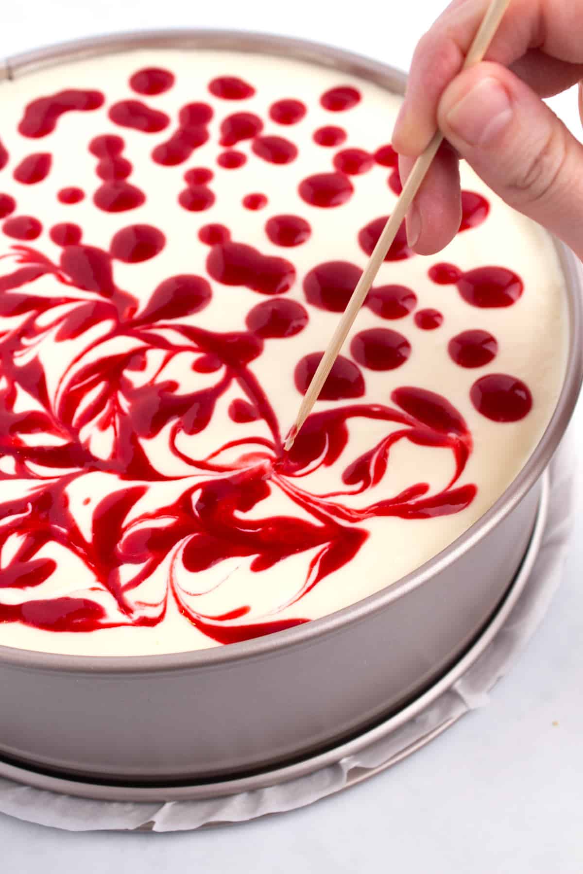 A toothpick swirling drops of raspberry topping into the surface of an unbaked cheesecake.