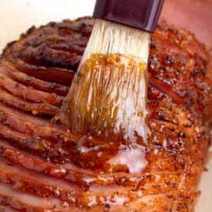 A brush saturated with glaze coating a large ham.